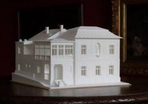 3 D model Vily Dominika Skuteckého vyhotovený z bieleho plastického materiálu. Je položený na stole. Na fotografii je zachytené priečelie budovy od hlavného vchodu. Budova je dvojpodlažná, obdĺžnikového tvaru. 