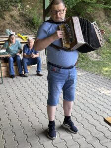 Vysoký muž stojí s harmonikou v náručí, na ktorej hrá